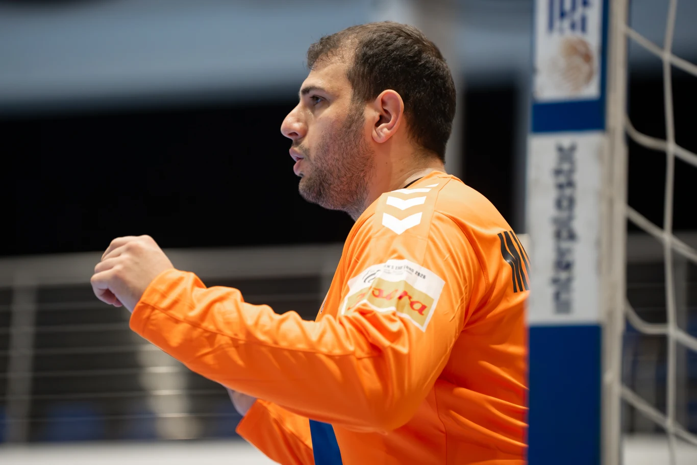 Bramkarz w pomarańczowym stroju sportowym stoi przy bramce podczas meczu piłki ręcznej, z wyraźnym skupieniem na twarzy i uniesionymi dłońmi gotowymi do obrony.