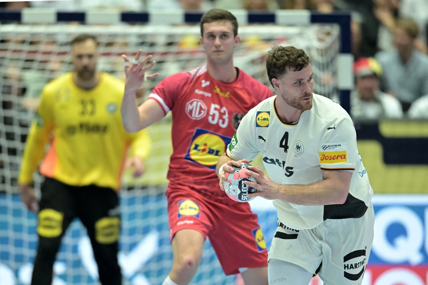 Dwóch zawodników piłki ręcznej w trakcie meczu, jeden ubrany w biały strój z piłką w rękach, drugi w czerwonym stroju próbujący go zatrzymać; w tle bramkarz i publiczność.