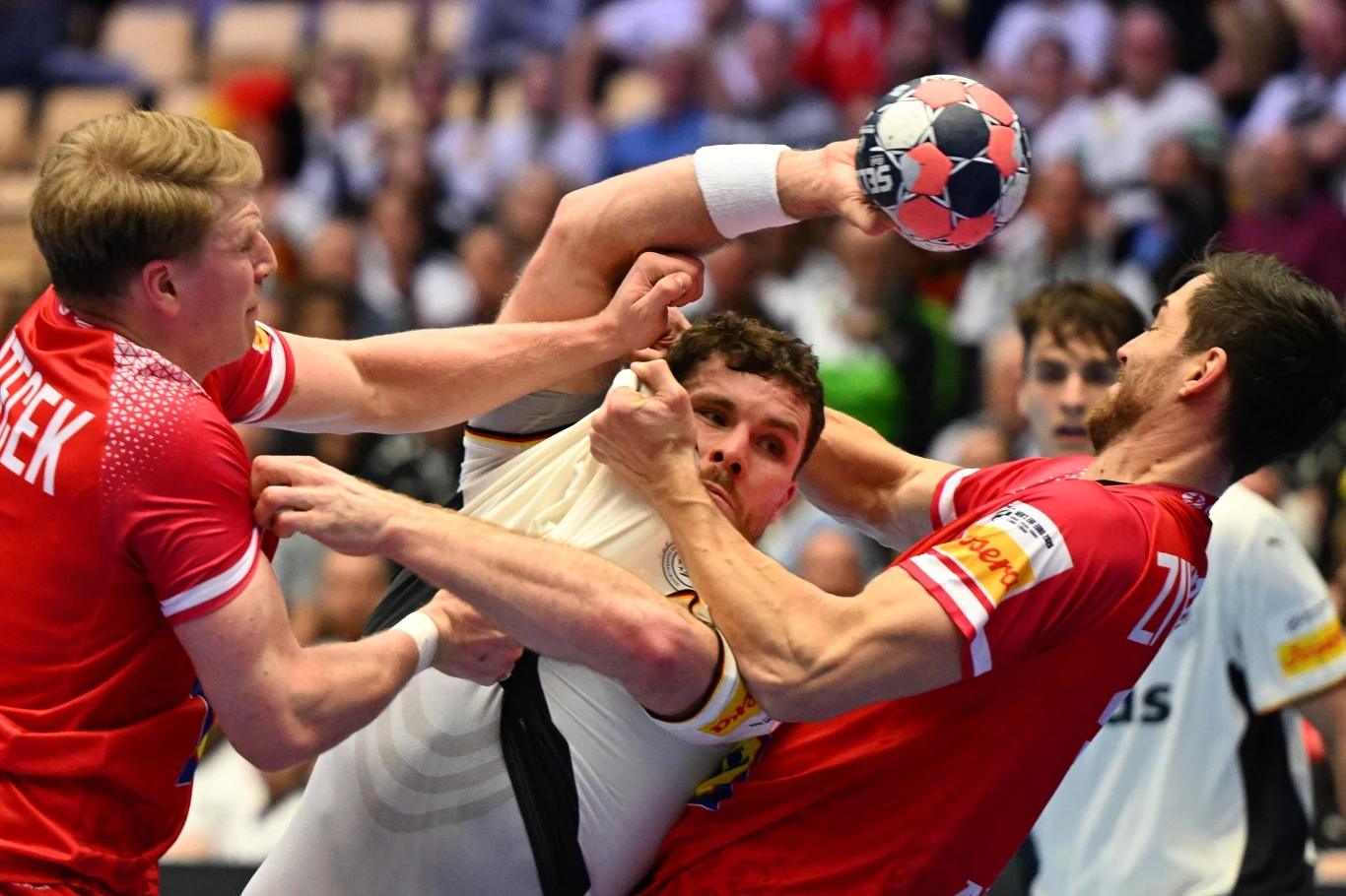 Dynamiczne starcie podczas meczu piłki ręcznej, trzech zawodników walczy o piłkę, dwóch w czerwonych koszulkach rywalizuje z jednym graczem w białym stroju, w tle rozmyta publiczność.