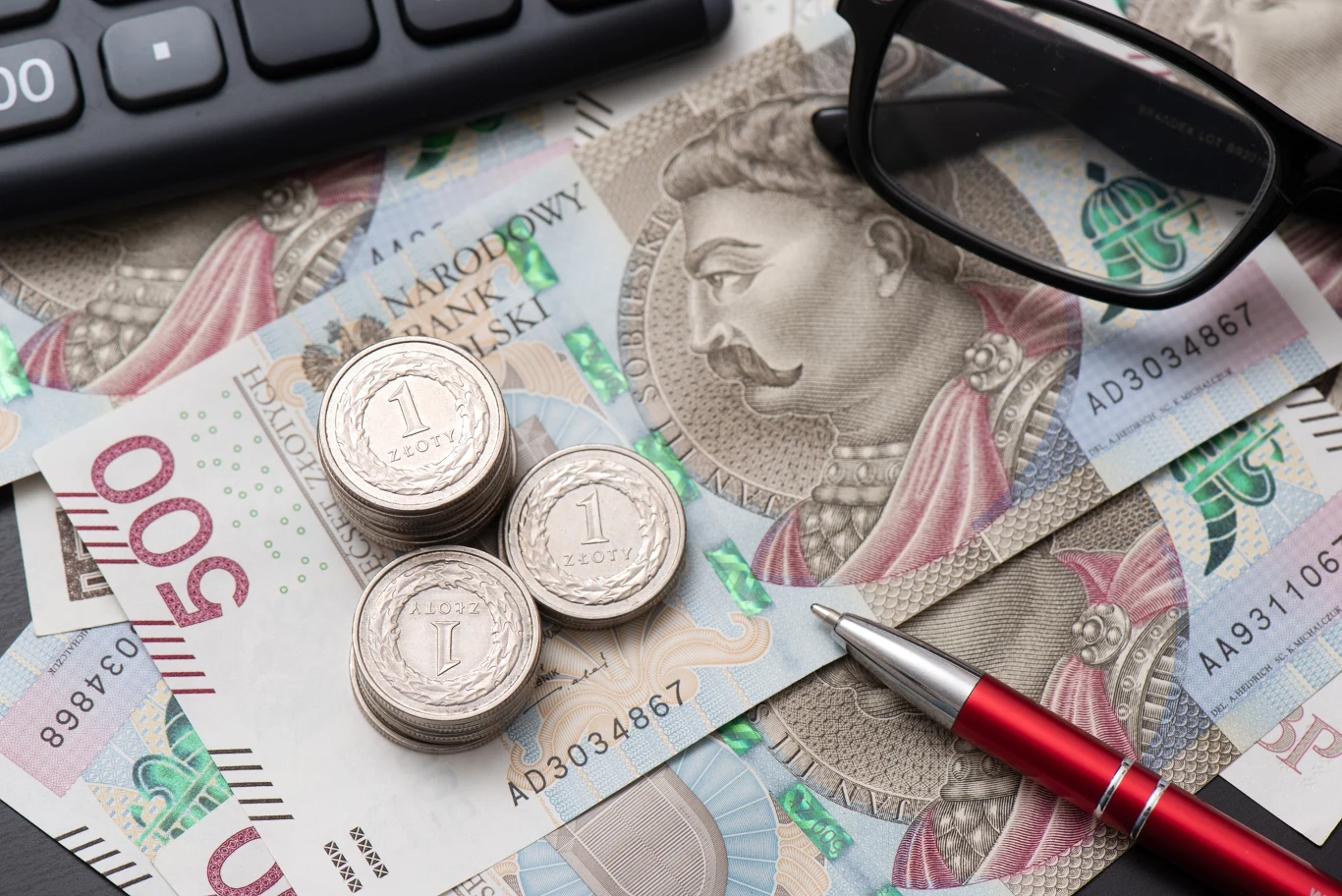 KSeF wchodzi w życie z początkiem lutego Stosy monet jednozłotowych ułożone na banknotach o nominale 500 złotych, w towarzystwie czerwonego długopisu, fragmentu klawiatury oraz okularów.