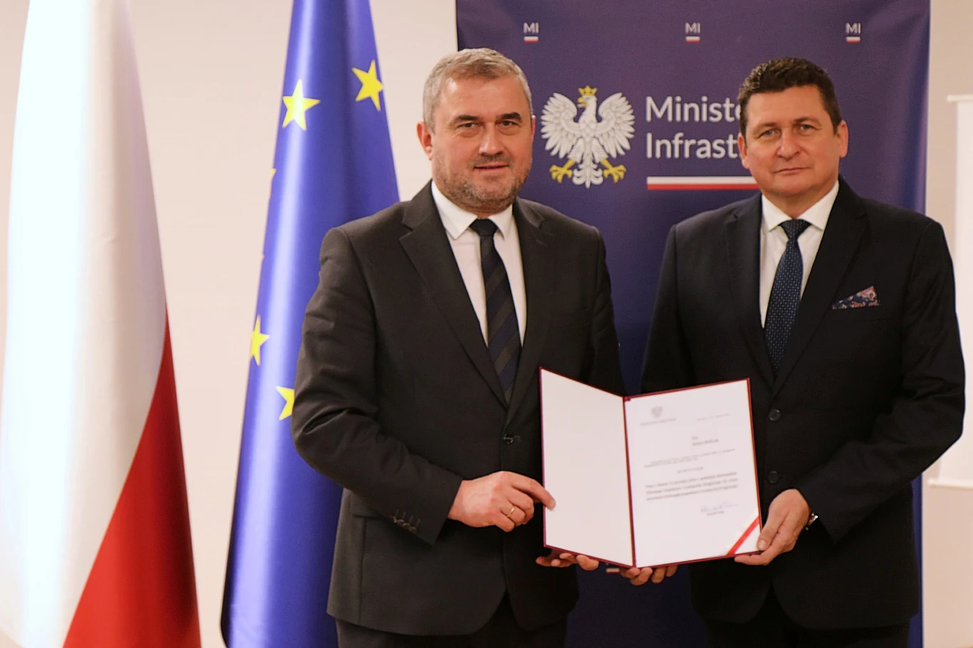 Dwóch mężczyzn w garniturach trzyma oficjalny dokument, w tle znajdują się flagi Polski oraz Unii Europejskiej, a także tablica z logo Ministerstwa Infrastruktury.