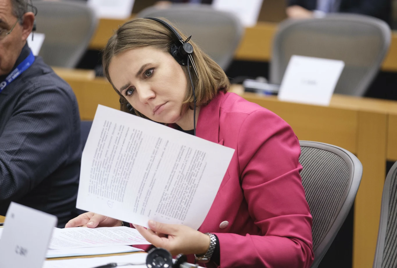 Kobieta w różowej marynarce z zestawem słuchawkowym na uszach uważnie przegląda dokumenty podczas spotkania lub konferencji, siedząc przy biurku w sali obrad.