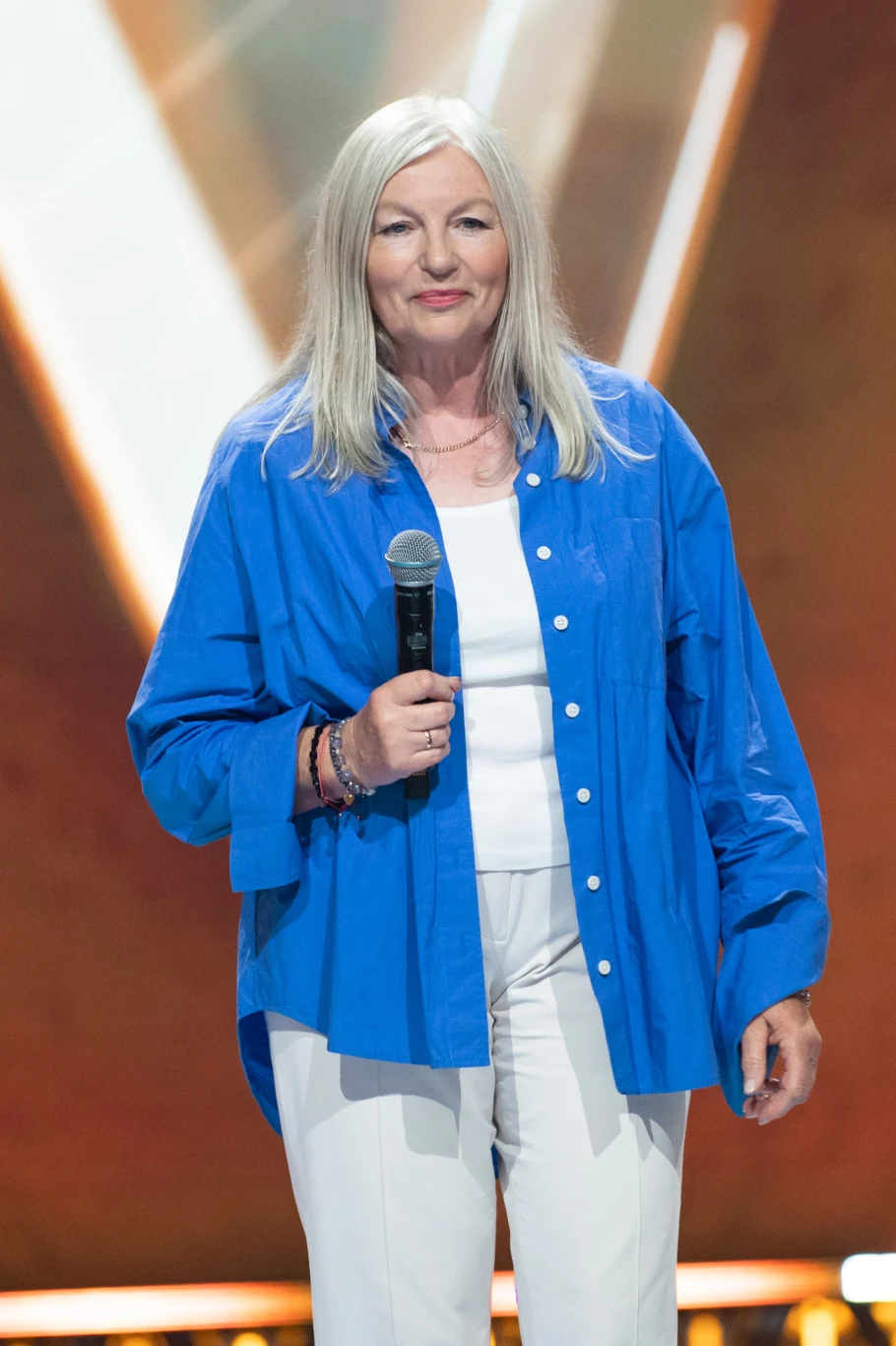 Kobieta o siwych, półdługich włosach w niebieskiej koszuli i białych spodniach trzyma mikrofon, stojąc na scenie, tło rozświetlone reflektorami z brązowymi tonacjami.