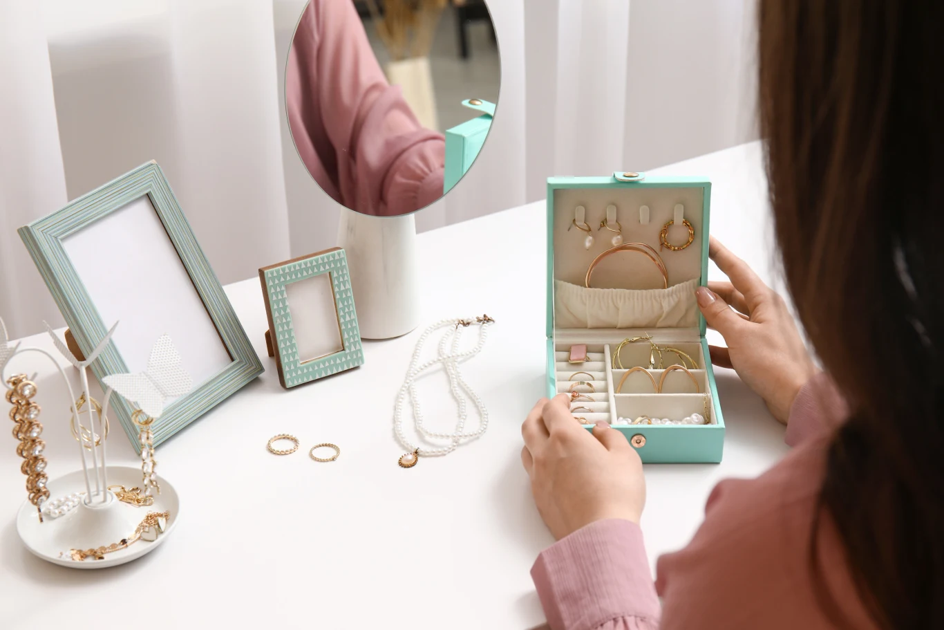 Zbliżenie kobiety siedzącej przy toaletce, która otwiera turkusowe pudełko z biżuterią. Na blacie stoją ramki na zdjęcia i okrągłe lustro, obok leżą naszyjnik, pierścionki i ekspozytor na bransoletki oraz kolczyki.