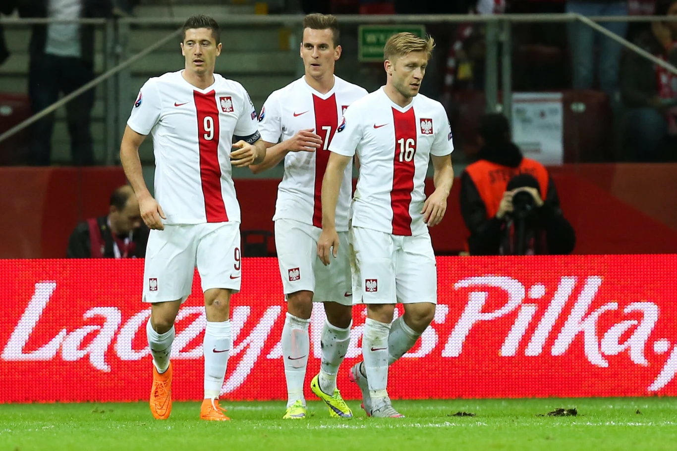 Trzech piłkarzy reprezentacji Polski w białych strojach z czerwonymi elementami stoi na murawie stadionu podczas meczu piłkarskiego. W tle widoczne są trybuny i reklama z napisem 'Łączy nas piłka'.
