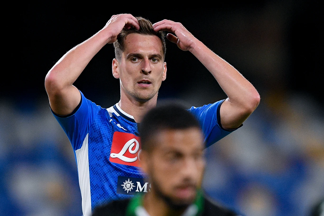 Piłkarz w niebieskiej koszulce drużyny SSC Napoli z widocznym logotypem sponsorów trzyma ręce na głowie w geście rozczarowania, na drugim planie nieostro widoczny zawodnik przeciwny, tło rozmyte z jasnymi światłami stadionu.