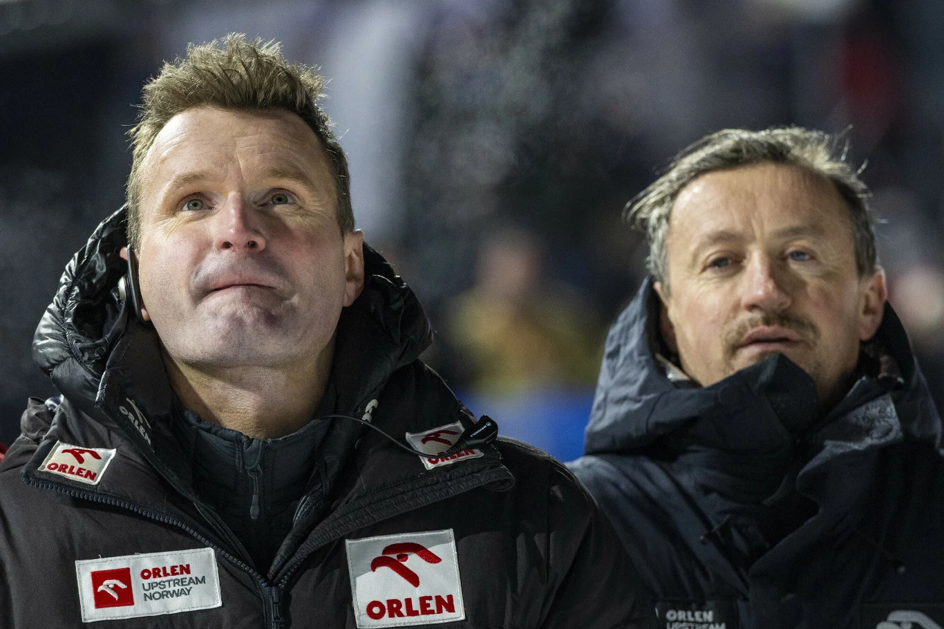 Dwóch mężczyzn w ciepłych kurtkach ze sponsorami Orlen, stojących blisko siebie, skupieni, w tle rozmyte światła.