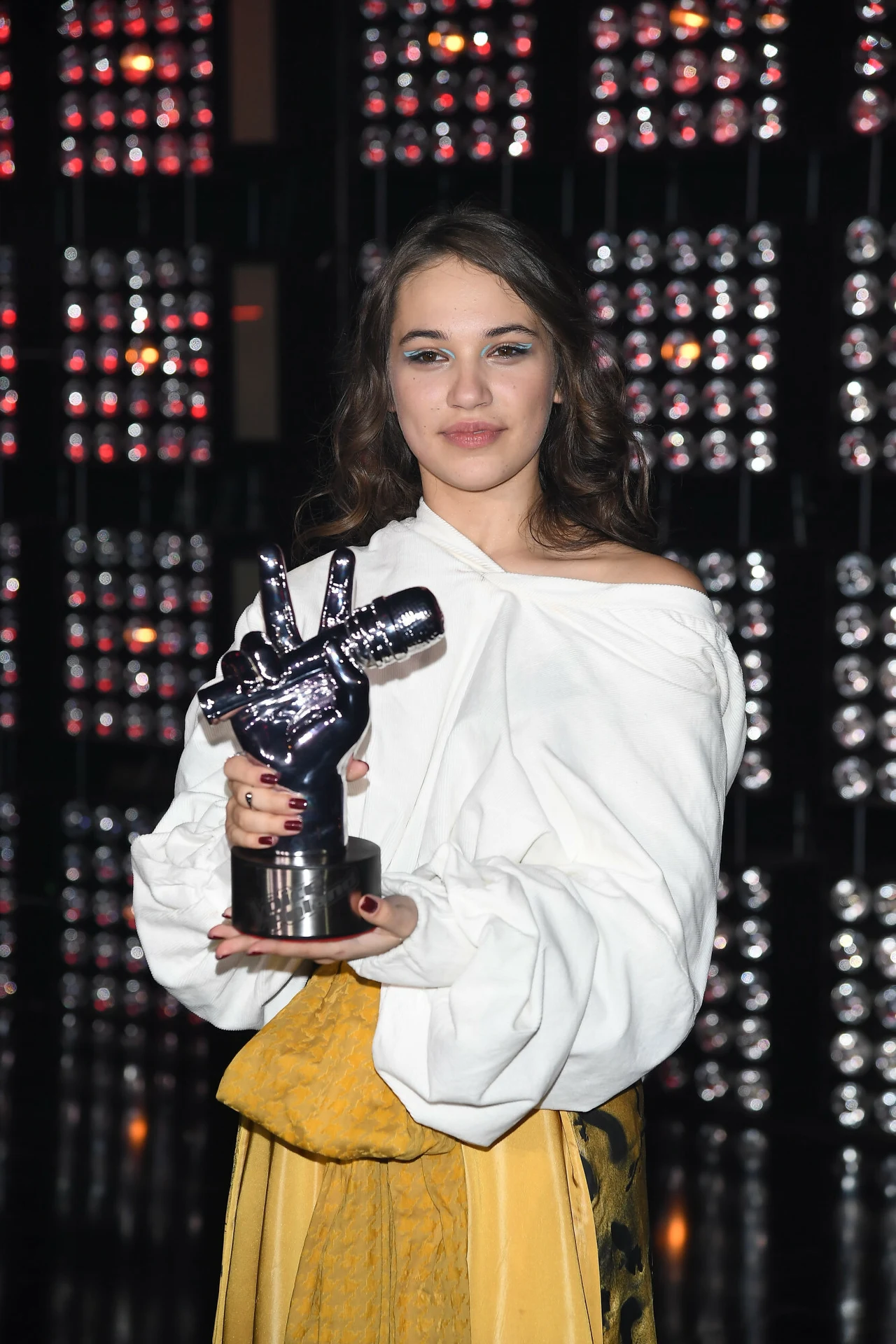 Młoda kobieta z długimi włosami, ubrana w białą bluzkę z odkrytym ramieniem oraz żółtą spódnicą, trzyma trofeum w kształcie dłoni wykonującej gest zwycięstwa, w tle ściana z połyskującymi światłami.