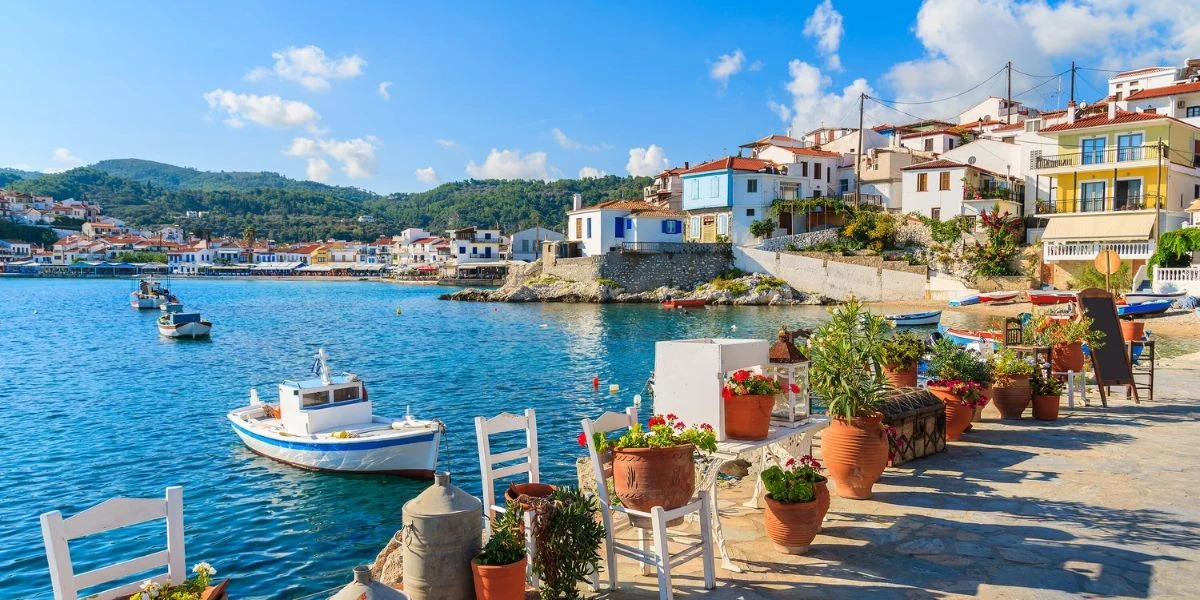 Nadmorski port z zacumowanymi łodziami, kolorowe domy na zboczu wzgórza, stoliki i krzesła ustawione przy brzegu oraz doniczki z kwiatami tworzą spokojną, śródziemnomorską atmosferę.