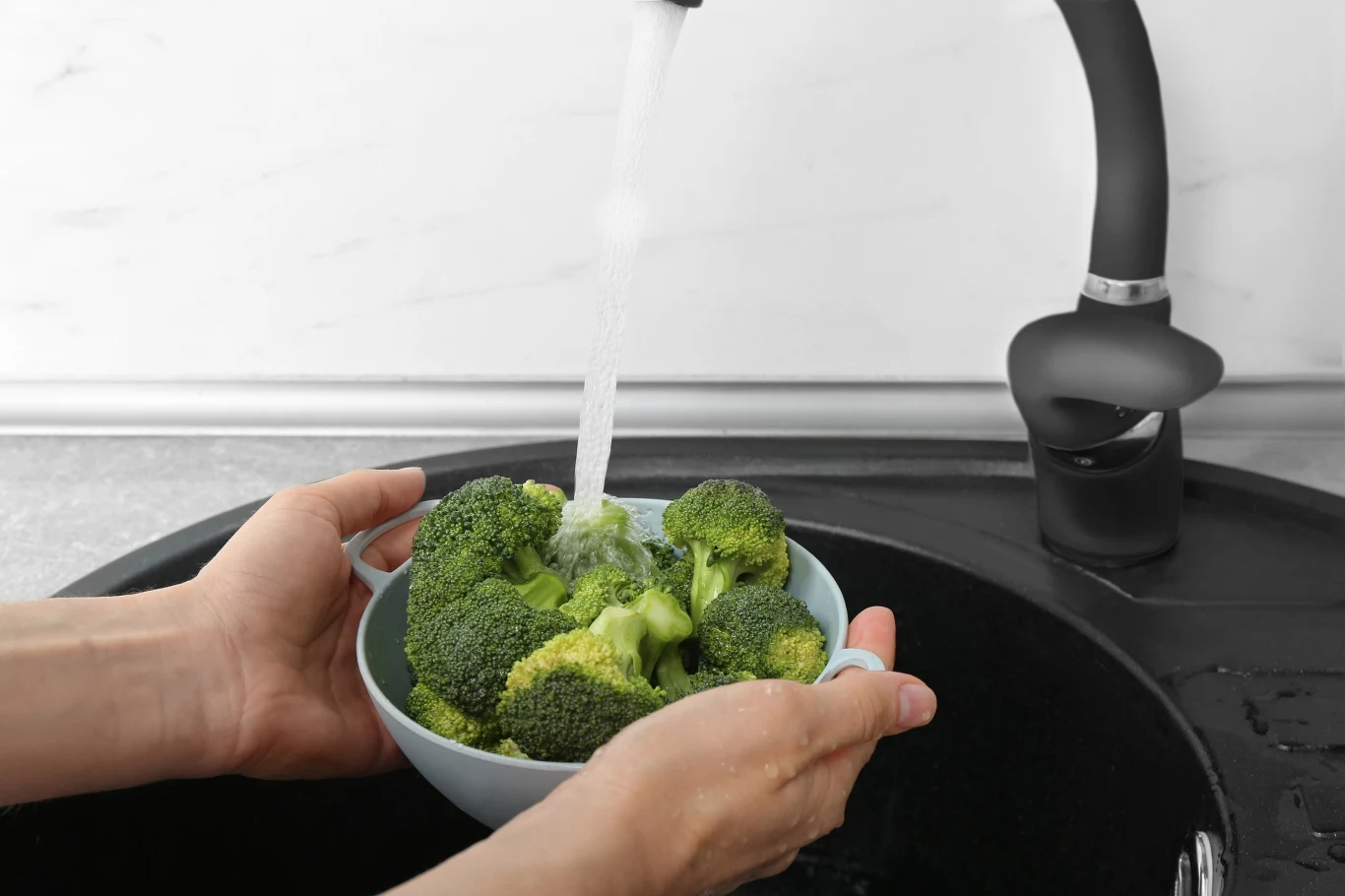 Miska pełna brokułów trzymana nad zlewem, na które leci strumień wody z kranu, co sugeruje mycie warzyw przed przygotowaniem posiłku.