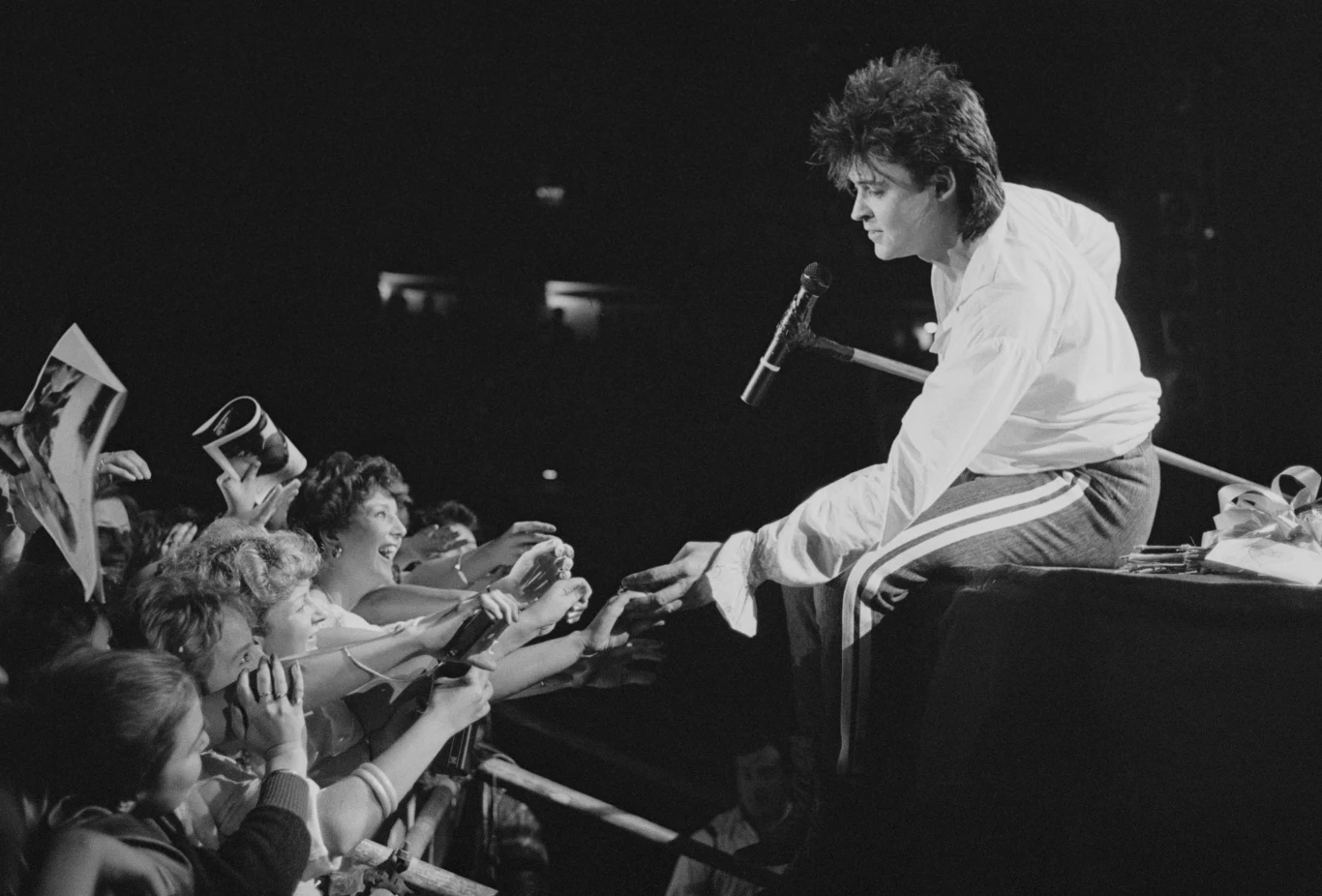 Wokalista na scenie nachyla się w stronę rozemocjonowanych fanów zgromadzonych przy barierce, podając rękę jednej z osób, podczas gdy inni wyciągają w jego stronę dłonie i trzymają plakaty.