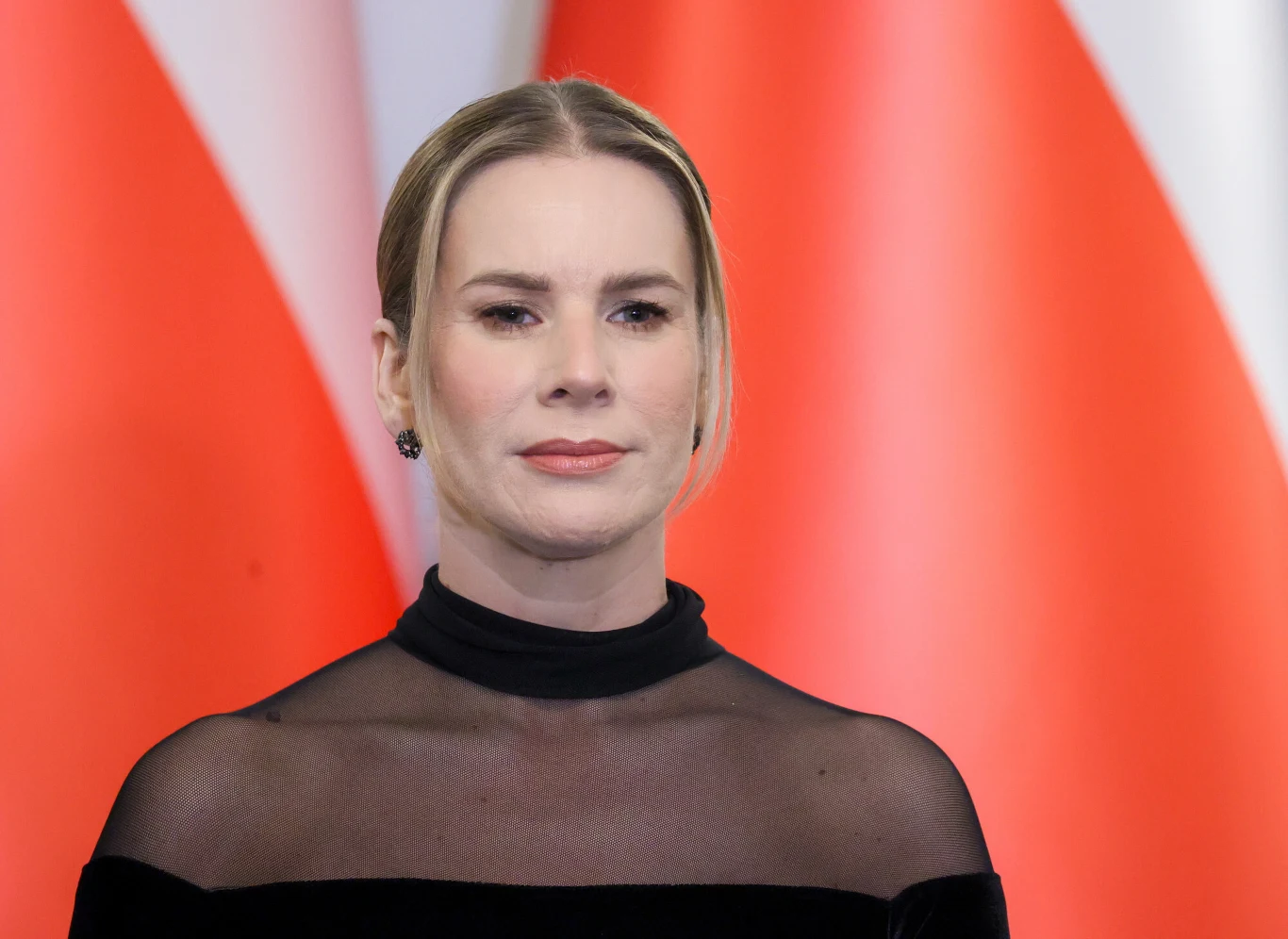 Marta Nawrocka w welurowej sukni Kobieta o jasnych włosach spiętych z tyłu ubrana w czarną suknię z przezroczystą górą stoi na tle dużych, rozmytych flag w czerwono-białych barwach.