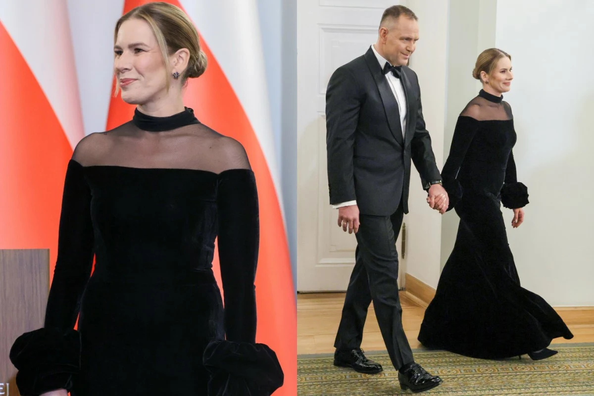 Pałac Prezydencki. Spotkanie noworoczne Pary Prezydenckiej z Korpusem Dyplomatycznym Kobieta w czarnej, długiej sukni z przezroczystą górą, z upiętymi włosami, stoi na tle biało-czerwonych flag. Obok mężczyzna w czarnym smokingu, trzymający ją za rękę, wspólnie idą przez wnętrze budynku.