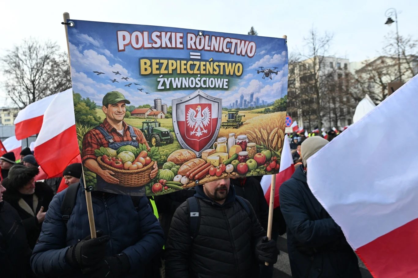 Protestujący trzymają transparent promujący polskie rolnictwo i bezpieczeństwo żywnościowe, otoczeni flagami Polski, z ilustracją rolnika oraz plonów rolnych na tle wiejskiego krajobrazu.