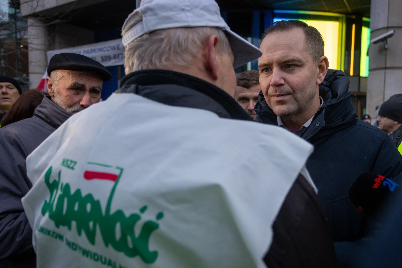 Kilka osób zgromadzonych na ulicy, mężczyzna w pierwszym planie ubrany w kamizelkę z napisem Solidarność rozmawia z innym mężczyzną. W tle budynek z jasnym oświetleniem oraz kolejni uczestnicy spotkania.