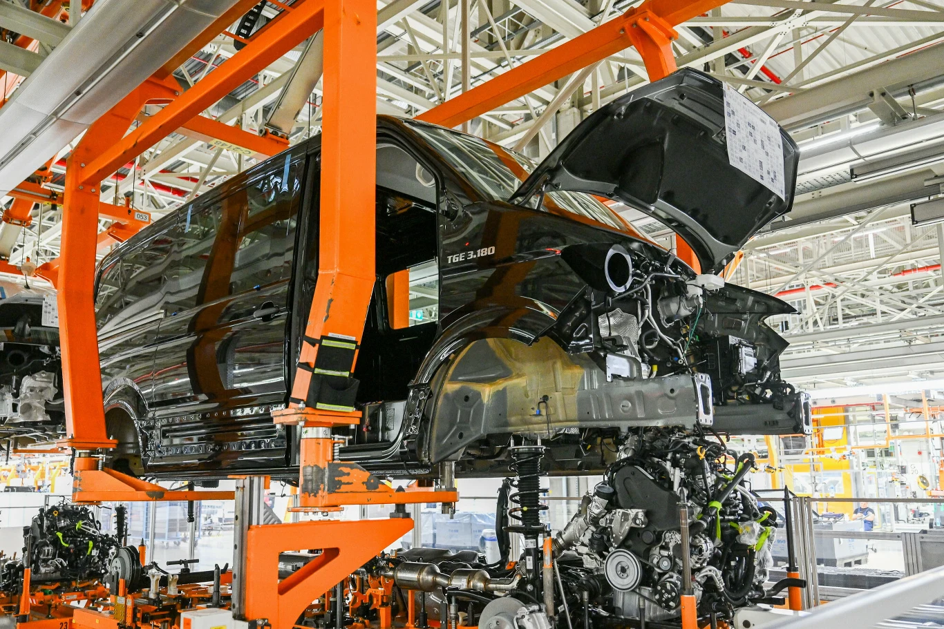 Czarna karoseria samochodu dostawczego zawieszona na pomarańczowej ramie w hali montażowej fabryki samochodów, widoczne części układu napędowego i elementy konstrukcyjne podwozia, wokół stalowa konstrukcja hali przemysłowej.