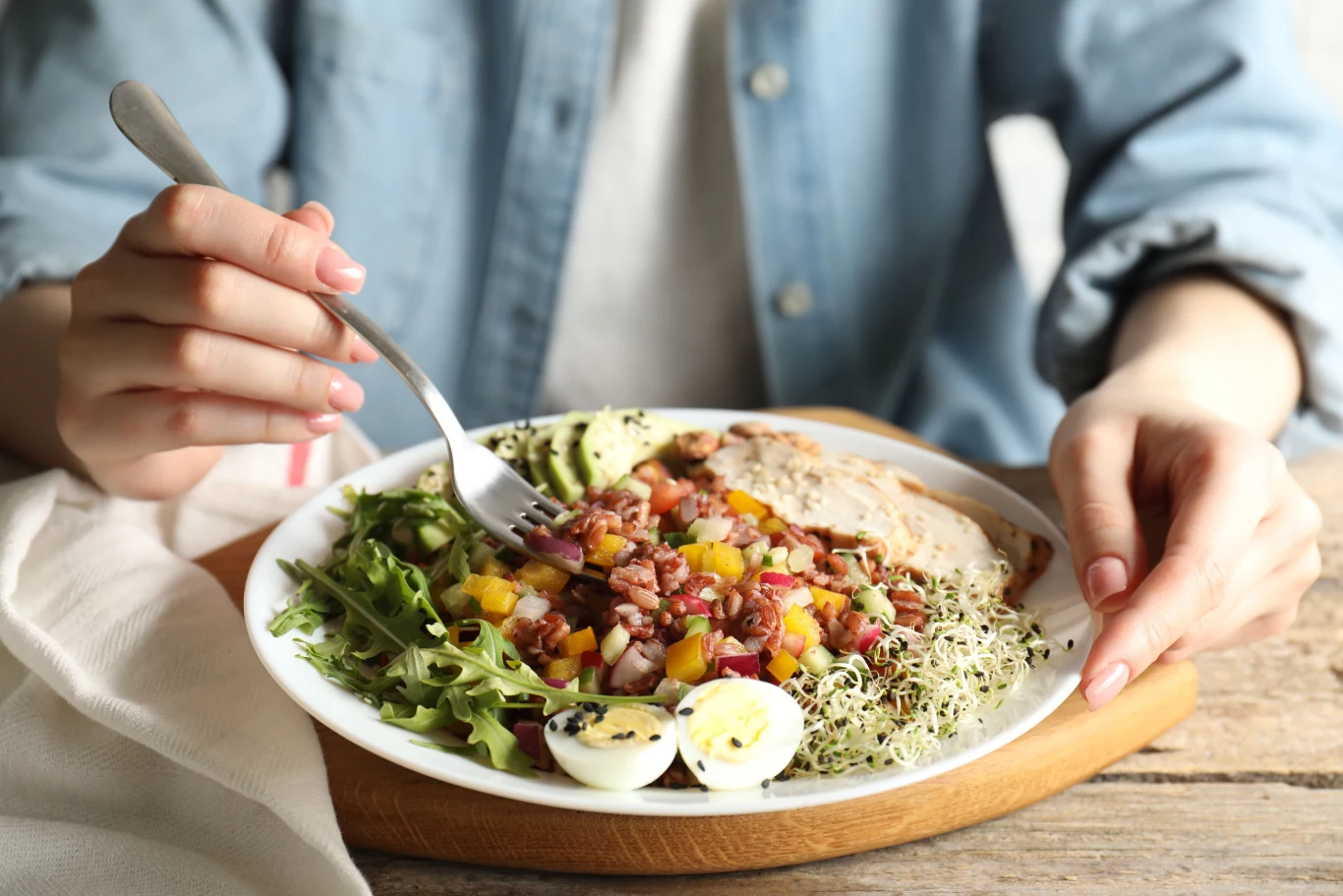 Jedzą je osoby z najzdrowszym sercem na świecie. Białko roślinne chroni przed chorobami Kolorowy talerz z sałatką składającą się z rukoli, kiełków, plasterków kurczaka, jajek przepiórczych, połówki awokado oraz warzyw pokrojonych w kostkę. Kobieta w niebieskiej koszuli trzyma widelec i zaczyna jeść.