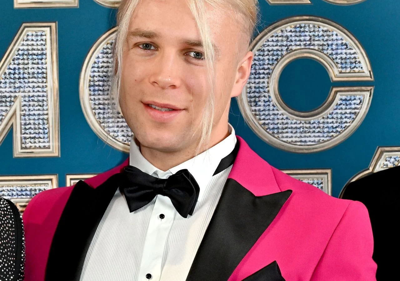 Skolim Mężczyzna z długimi blond włosami uczesanymi na bok, ubrany w różową marynarkę, białą koszulę i czarną muchę, stoi na tle dekoracyjnej ściany z dużymi srebrnymi literami.