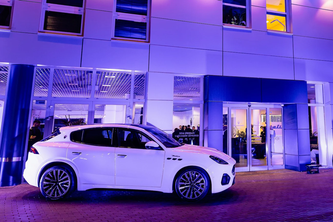 Luksusowy biały samochód terenowy zaparkowany przed nowoczesnym budynkiem oświetlonym fioletowym światłem, za szybami widoczni są ludzie uczestniczący w wydarzeniu wewnątrz.