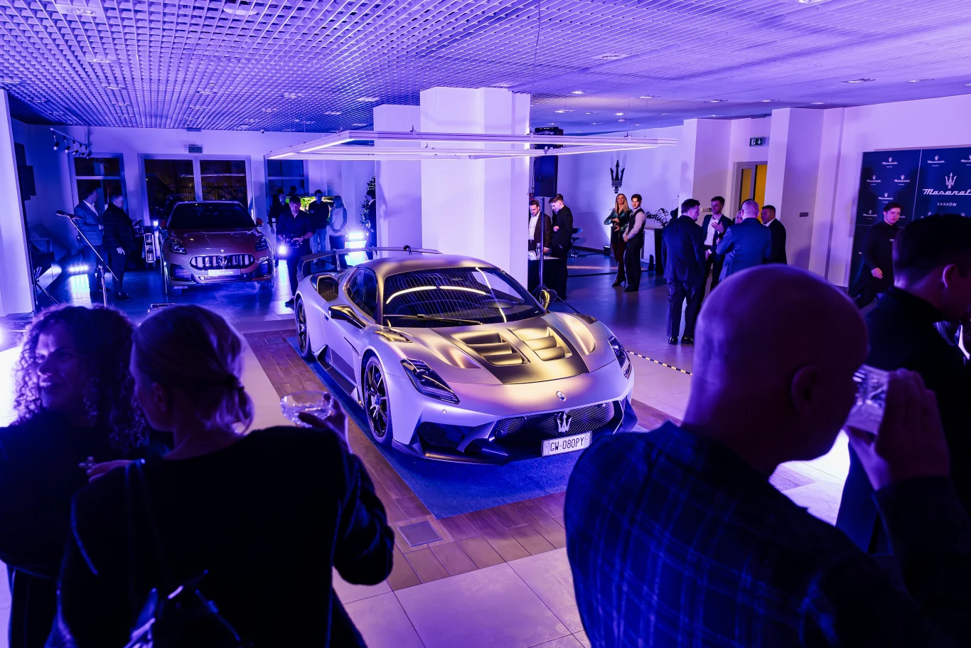 Luksusowy samochód sportowy Maserati MC20 prezentowany w salonie samochodowym podczas eventu, otoczony przez osoby w eleganckich strojach, podświetlony niebieskim światłem, w tle widoczne inne auta oraz uczestnicy wydarzenia rozmawiający w grupach.