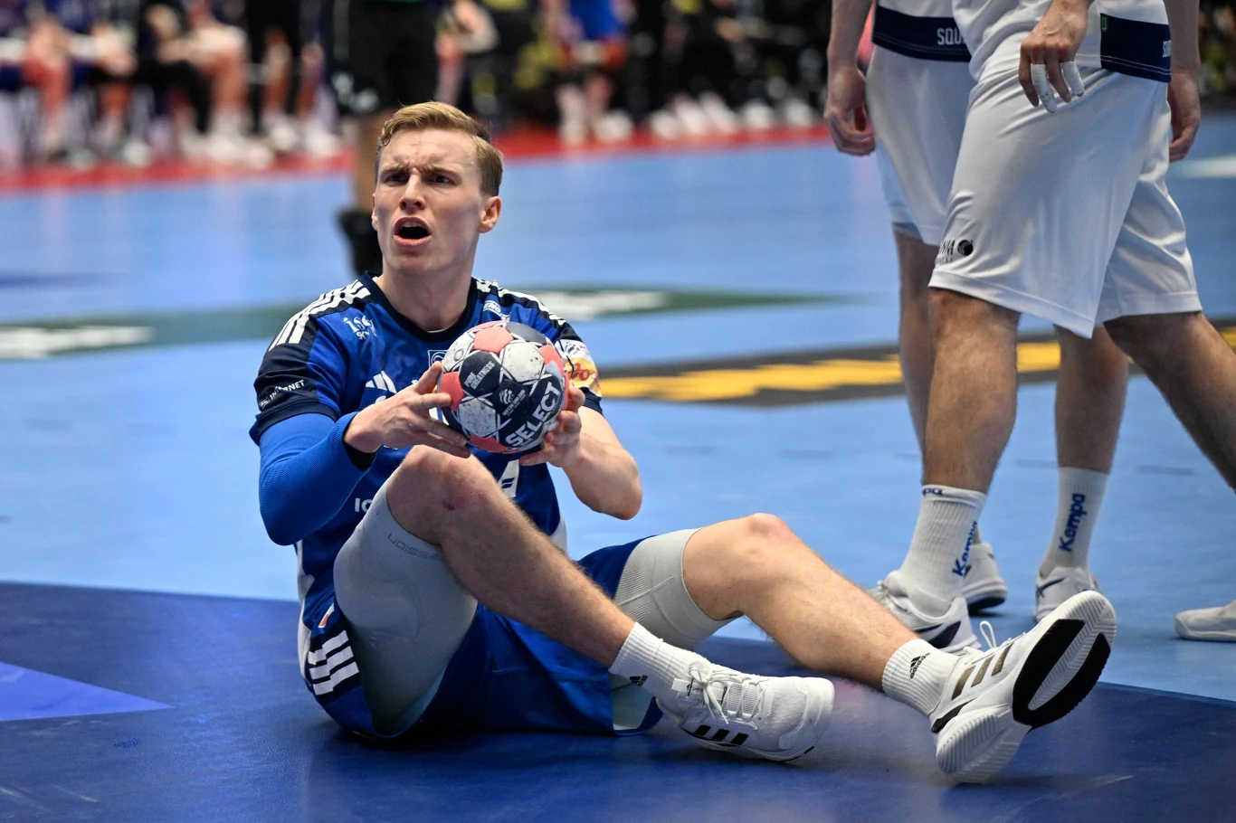 Zawodnik w niebieskim stroju siedzi na parkiecie trzymając piłkę ręczną z wyrazem zdziwienia lub frustracji na twarzy. Wokół widoczne są nogi innych zawodników oraz fragment boiska sportowego.