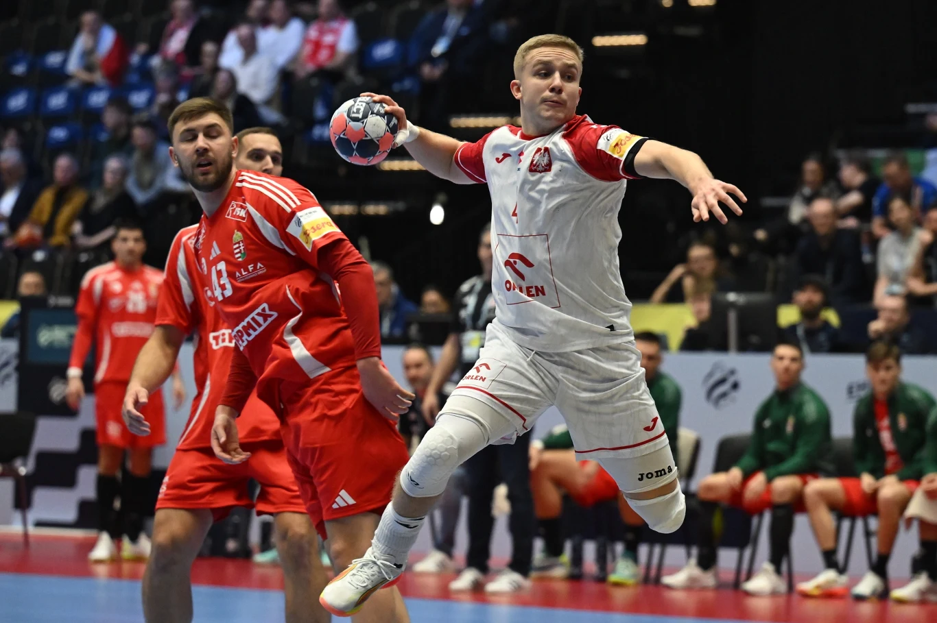 Zawodnik w białym stroju podczas skoku oddaje rzut piłką ręczną w trakcie meczu, wokół niego gracze drużyny przeciwnej w czerwonych strojach, na trybunach widoczna publiczność.