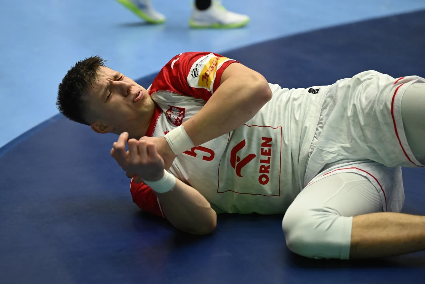 Zawodnik w stroju sportowym drużyny narodowej Polski leży na parkiecie trzymając się za rękę, mając wyraz bólu na twarzy. Na koszulce widoczne logo sponsora. W tle fragment boiska i inne rozeznawalne elementy rozgrywek sportowych.
