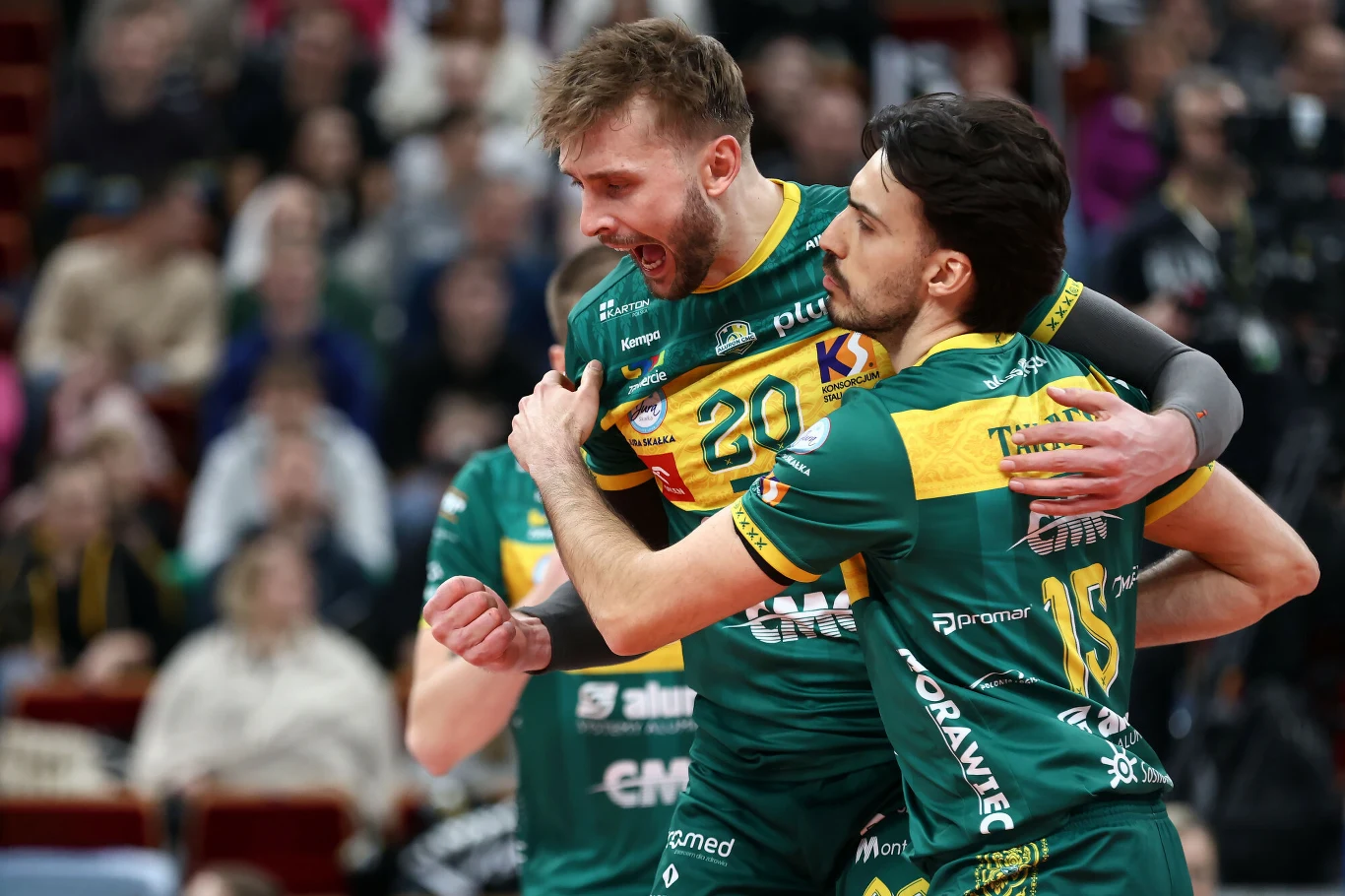 Dwóch siatkarzy w zielonych strojach drużynowych podczas intensywnej celebracji punktu na tle rozmytej publiczności. Jeden z nich z uniesioną ręką i wyrazistą ekspresją twarzy, drugi obejmuje go ramieniem.