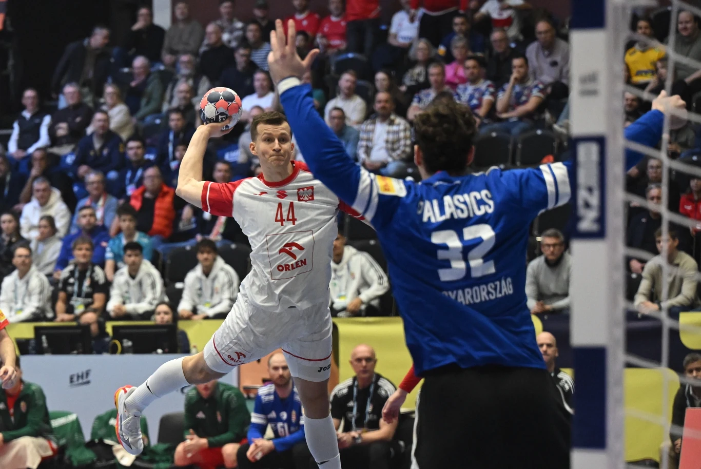Polski szczypiornista w białym stroju wykonuje rzut w kierunku bramki strzeżonej przez węgierskiego bramkarza ubranego na niebiesko, na tle publiczności kibicującej z trybun.
