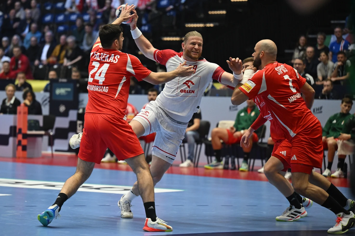 Zawodnik w białym stroju próbujący przedrzeć się przez dwóch obrońców w czerwonych koszulkach podczas meczu piłki ręcznej, widoczne dynamiczne starcie oraz wyraziste emocje sportowców.