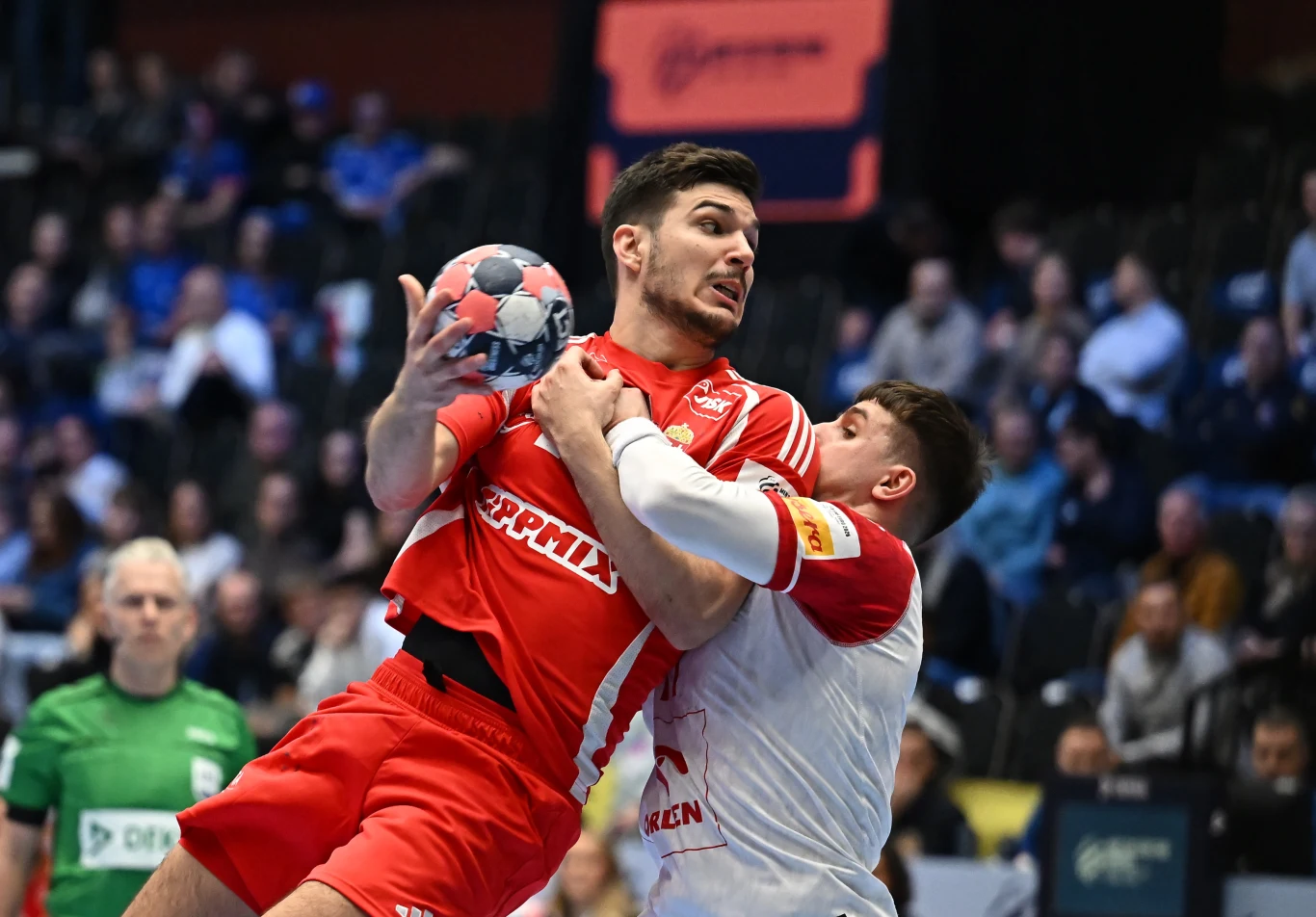 Dwóch zawodników walczy zaciekle o piłkę ręczną na tle pełnych trybun kibiców, jeden z zawodników w czerwonym stroju unosi piłkę w górę, podczas gdy przeciwnik w białym stroju próbuje go zatrzymać.