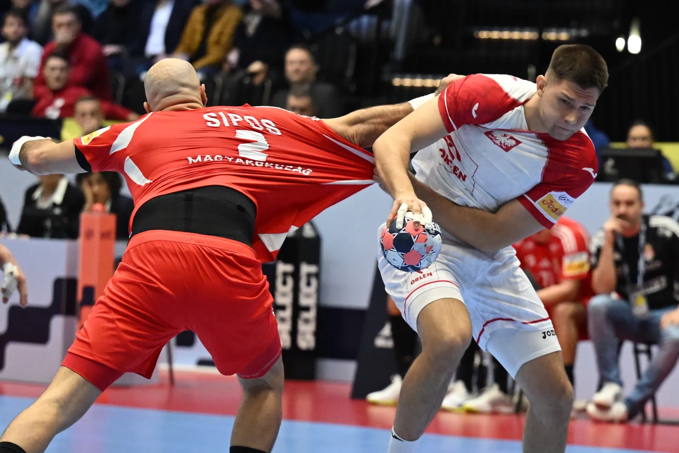 Dwóch zawodników w trakcie meczu piłki ręcznej, jeden z nich trzyma piłkę i jest silnie ciągnięty za koszulkę przez przeciwnika. Intensywna walka o piłkę na sportowej hali, w tle widoczni kibice.