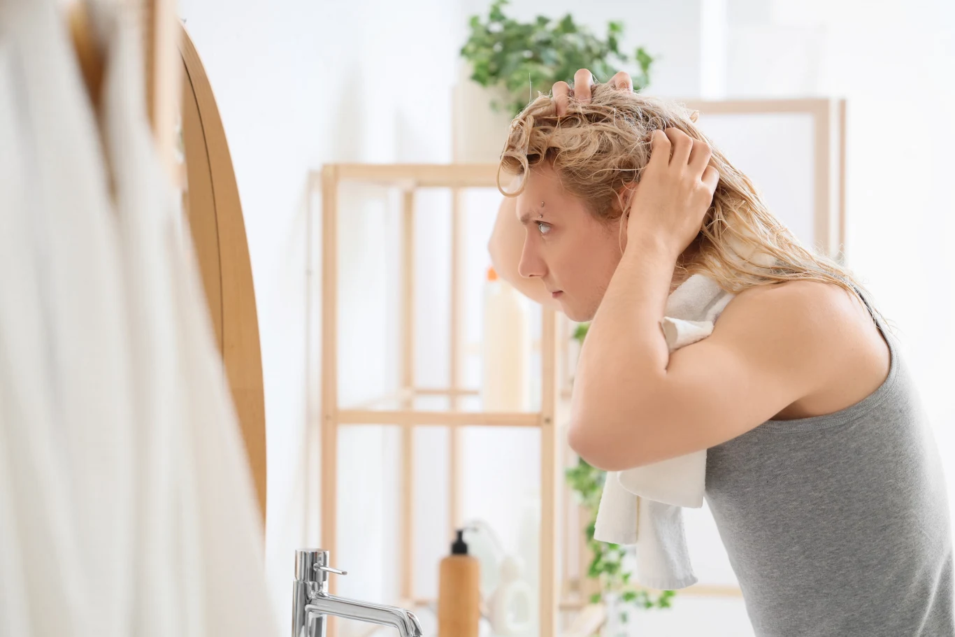 Kobieta stoi przy umywalce, suszy ręcznikiem mokre włosy. Na twarzy widać skupienie. W tle widać jasne, minimalistyczne wnętrze z elementami drewnianymi i zieloną rośliną.