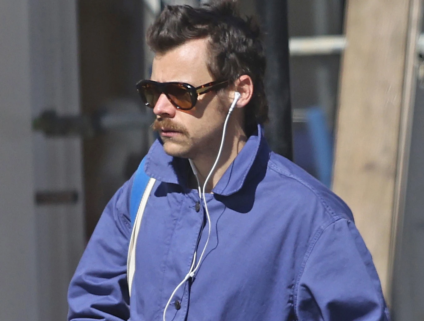 Harry Styles ujawnił pierwsze szczegóły nowej płyty Mężczyzna z ciemnymi okularami przeciwsłonecznymi i wąsem ubrany w niebieską kurtkę słucha muzyki przez słuchawki douszne, nosi biały pasek na ramieniu, stoi na zewnątrz podczas dnia.