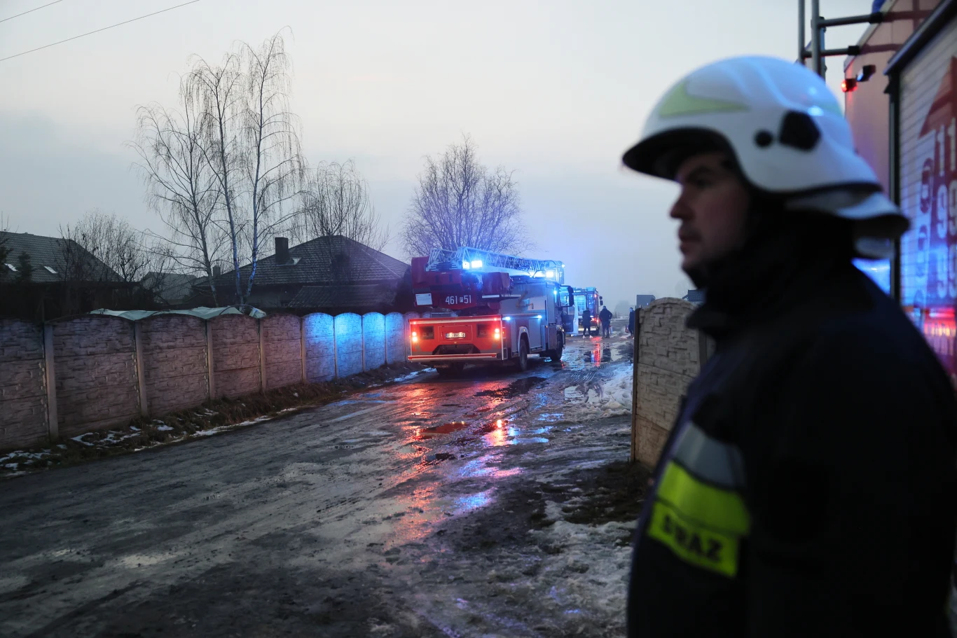 Pożar w Myjomicach. W szpitalu w Kępnie zmarła 38-latka, wcześniej zginął jej syn Strażak w mundurze i kasku na pierwszym planie, w tle dwa wozy strażackie z włączonymi światłami stojące na mokrej, zaśnieżonej ulicy w pobliżu ogrodzonych domów.