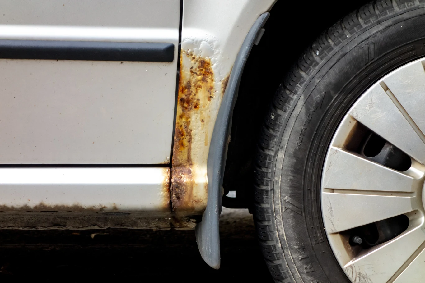 Fragment karoserii srebrnego samochodu osobowego z widocznym ogniskiem rdzy przy nadkolu i śladami korozji na progu, po prawej stronie koło z plastikowym kołpakiem.