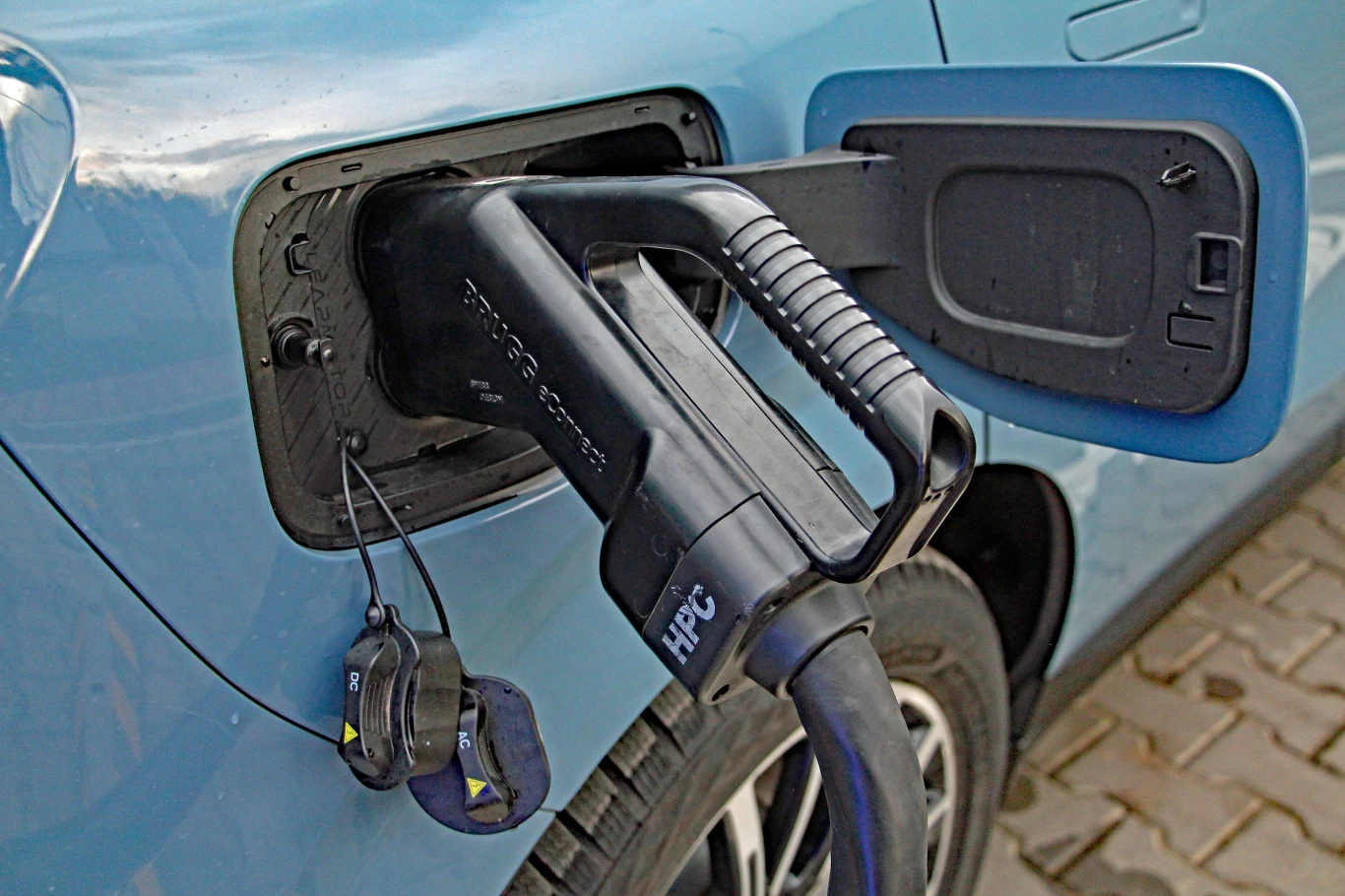 Wtyczka ładowarki elektrycznej podłączona do portu ładowania samochodu osobowego, niebieski lakier auta, widoczna kostka brukowa pod kołami.