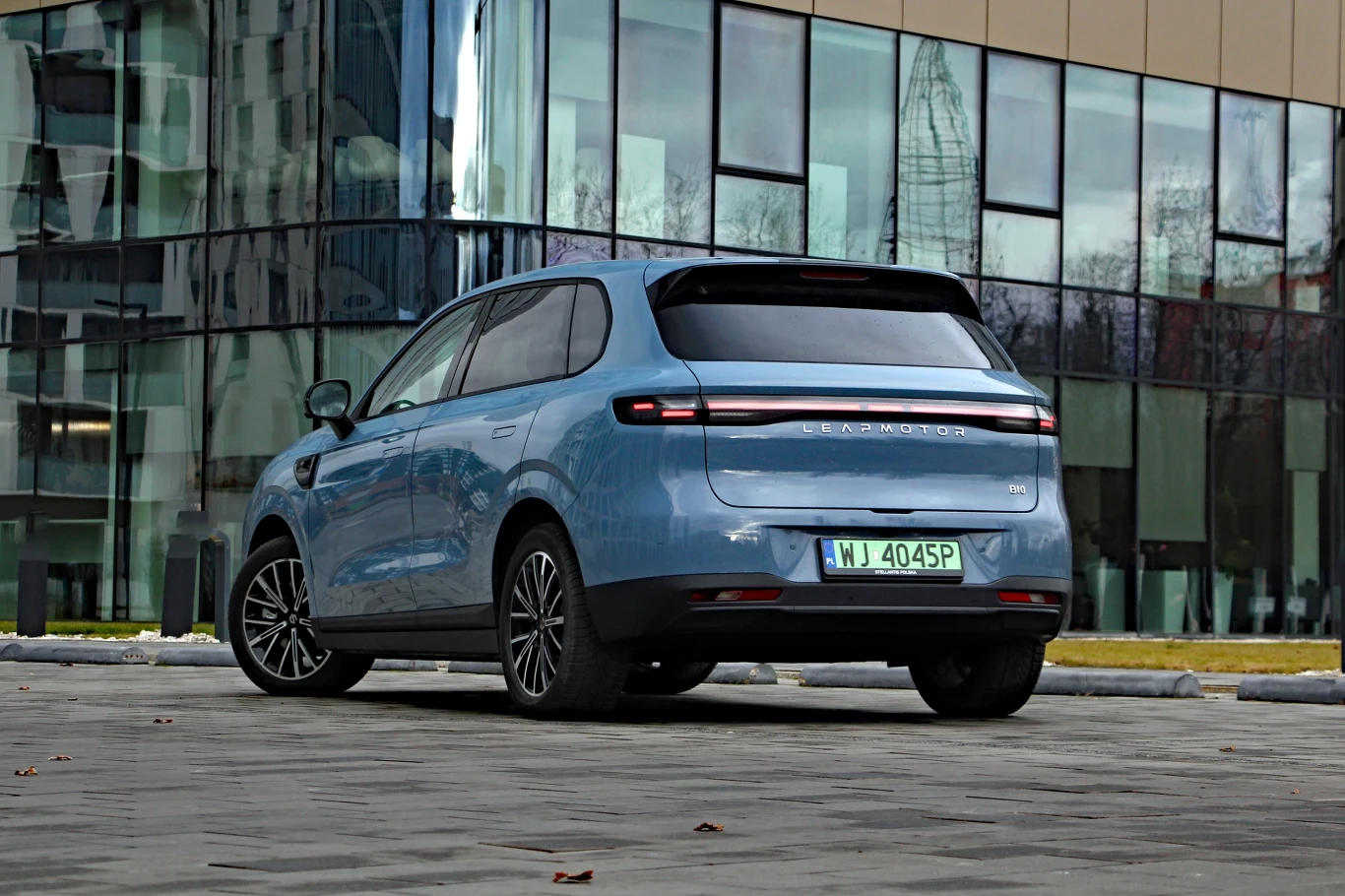 Jasnoniebieski samochód osobowy ustawiony tyłem na brukowanym placu przed nowoczesnym przeszklonym budynkiem, w tle odbicia drzew i wieżowców w szybach.