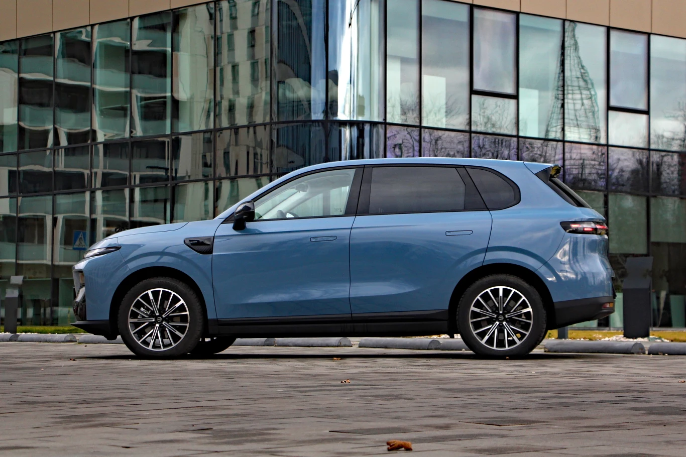 Nowoczesny, niebieski SUV zaparkowany na tle szklanej fasady nowoczesnego budynku. Lśniąca karoseria i czyste linie samochodu podkreślają miejski charakter scenerii, a lekko odbite sylwetki w oknach dodają dynamiki całości.