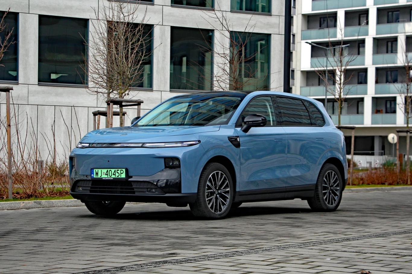 Niebieski samochód osobowy nowoczesnego typu SUV zaparkowany na brukowanym parkingu miejskim, w tle szare elewacje nowoczesnych budynków mieszkalnych i nagie zimowe drzewa.