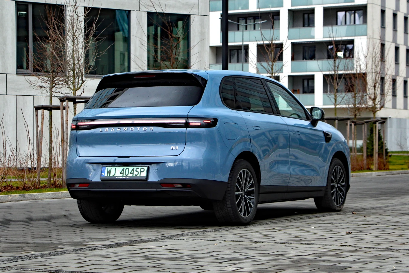 Niebieski samochód osobowy marki Seres 5 EV zaparkowany na kostce brukowej przy nowoczesnych budynkach mieszkalnych, otoczenie jest niemal pozbawione zieleni, w tle widoczne drzewa bez liści.