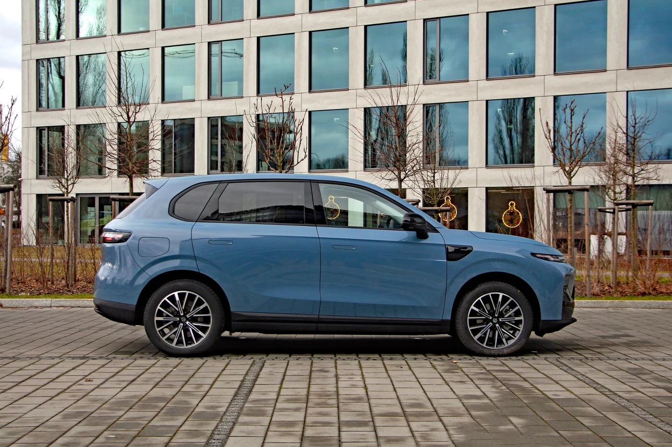 Niebieski samochód typu SUV zaparkowany na tle nowoczesnego budynku biurowego z dużymi oknami, w otoczeniu nagich drzew oraz brukowanego chodnika.