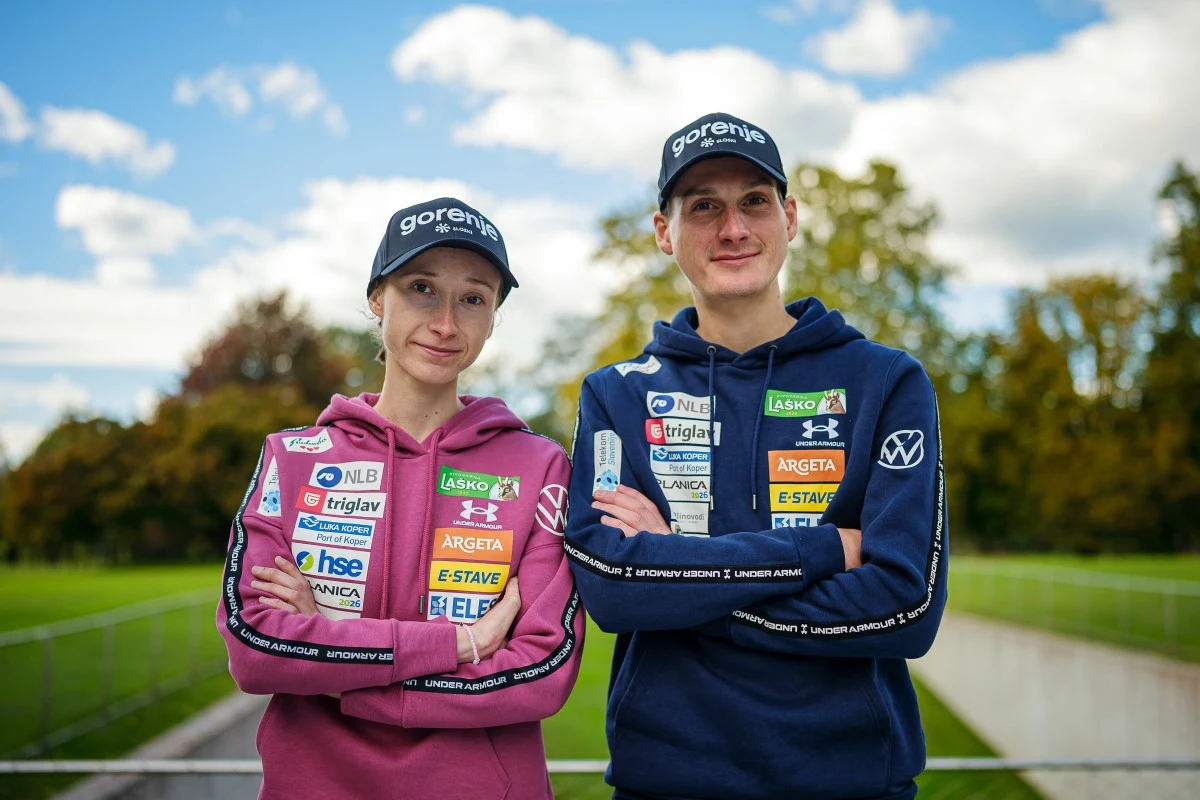 Dwie uśmiechnięte osoby stojące obok siebie na zewnątrz, noszące sportowe bluzy z licznymi logotypami sponsorów oraz czapki z daszkiem, w tle widoczna zieleń i niebieskie niebo z chmurami.