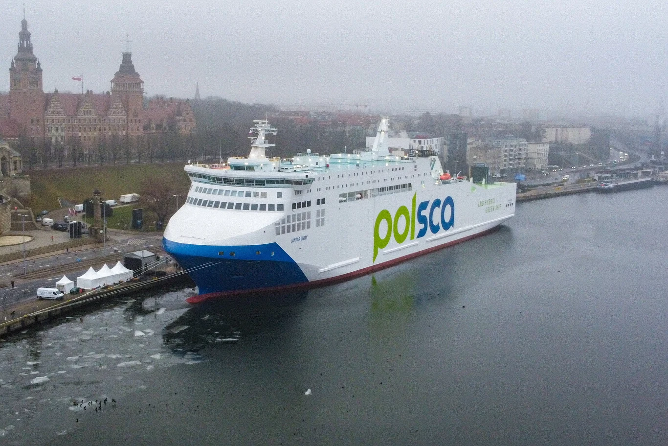 Szczecin. Nowy prom "Jantar" został zaprezentowany przy Wałach Chrobrego Duży nowoczesny prom pasażersko-towarowy oznaczony napisem 'Polska' zacumowany w miejskim porcie, po lewej stronie widoczna zabytkowa zabudowa z czerwonymi dachami, w tle rozciąga się zamglone miasto.