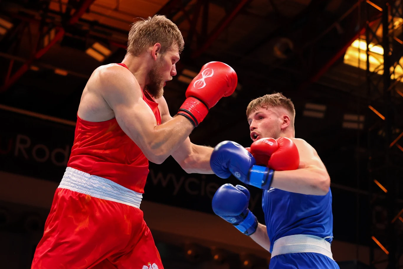 Dwóch bokserów w trakcie dynamicznej wymiany ciosów na ringu, jeden ubrany w strój czerwony, drugi w niebieski, światło i otoczenie hali sportowej podkreślają atmosferę rywalizacji.