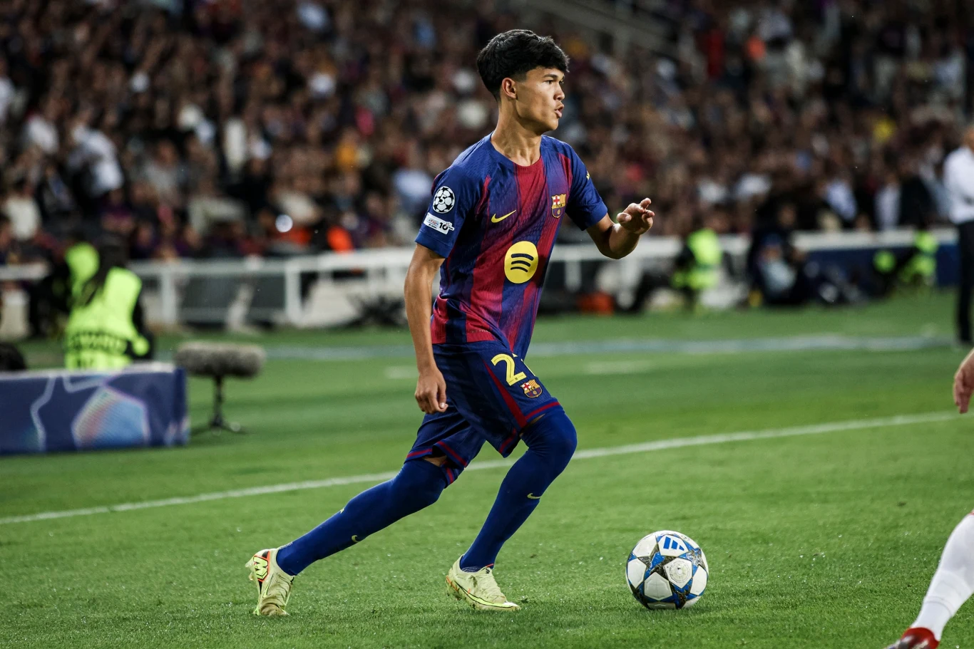 Piłkarz FC Barcelony w trakcie meczu, ubrany w strój drużynowy, prowadzi piłkę nożną po murawie stadionu przy pełnych trybunach i obecności fotoreporterów w tle.