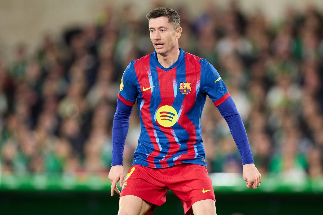 Piłkarz w stroju FC Barcelony podczas meczu piłkarskiego, skoncentrowany na grze, na tle rozmytego stadionu i kibiców.