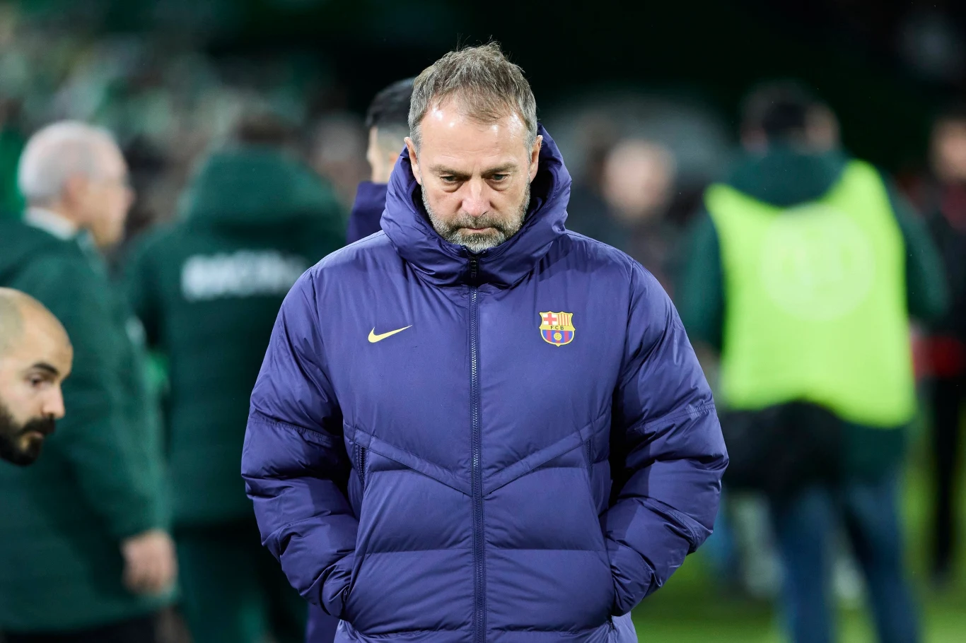 Mężczyzna w kurtce z herbem klubu FC Barcelona stoi na boisku z opuszczoną głową, w tle rozmyte postacie i zielone otoczenie stadionu.