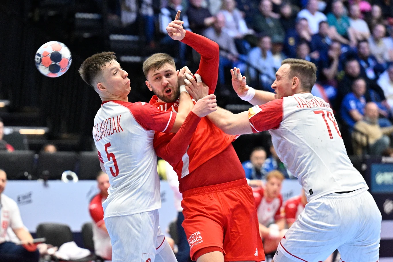 Dynamiczna walka podczas meczu piłki ręcznej, trzech zawodników w intensywnym starciu, dwóch ubranych w białe stroje próbuje powstrzymać gracza w czerwonym stroju, piłka znajduje się w powietrzu.