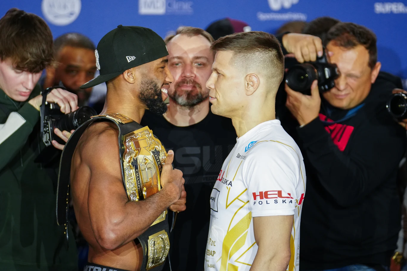 Dwaj zawodnicy mieszanych sztuk walki stoją naprzeciwko siebie twarzą w twarz podczas oficjalnej ceremonii ważenia, jeden z nich trzyma mistrzowski pas, otoczeni są przez fotoreporterów i kamerzystów.